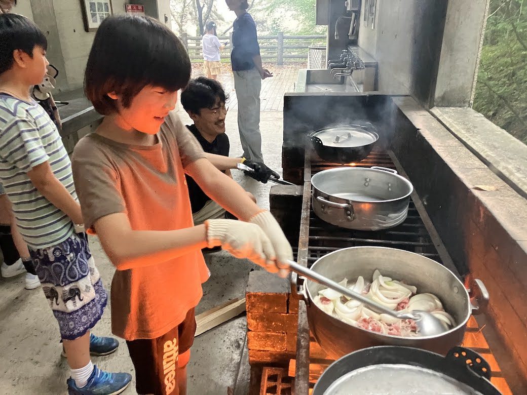修学旅行のかまど料理で、軍手をしてフライパンを混ぜる男の子。火おこしから調理まで子どもたちが取り組む場面