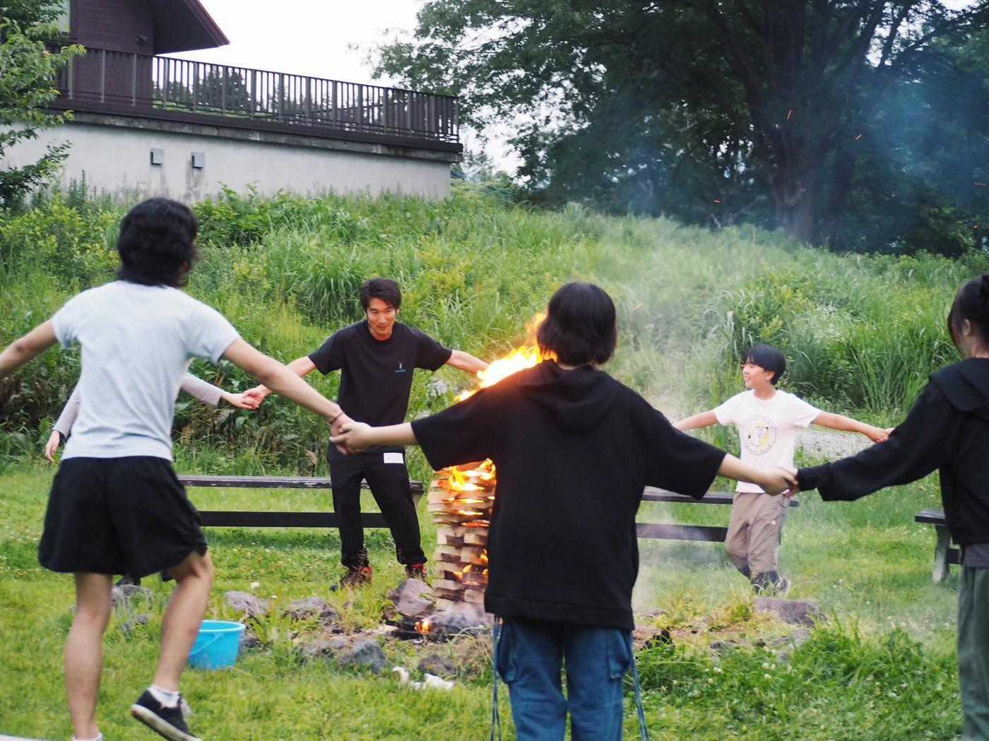 燃え上がるキャンプファイヤーを囲んで手をつなぎ踊るaini schoolの生徒とスタッフたち