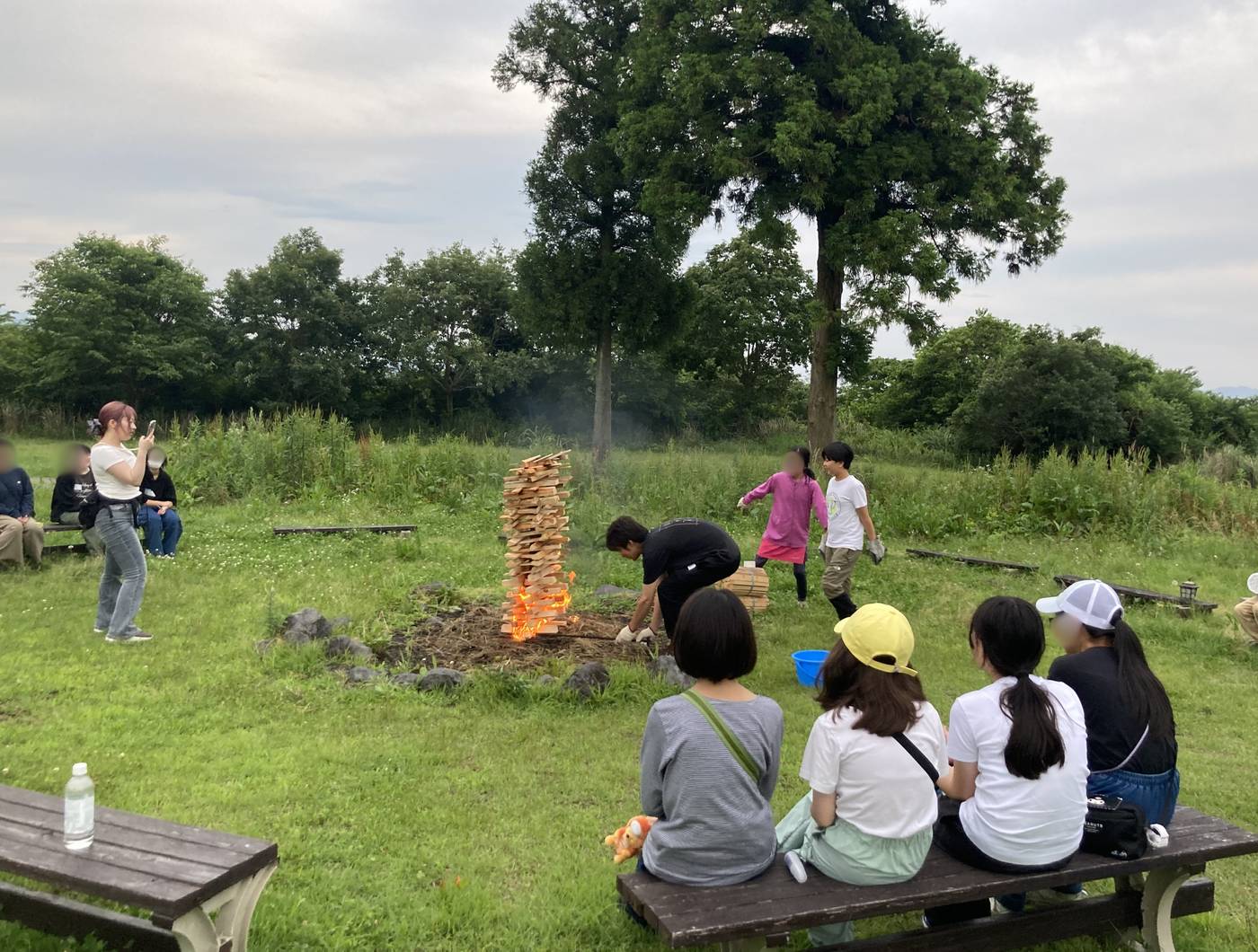  草原の広場で薪に点火し、燃え上がり始めたキャンプファイヤーをaini schoolの生徒・スタッフが囲んで見守る様子