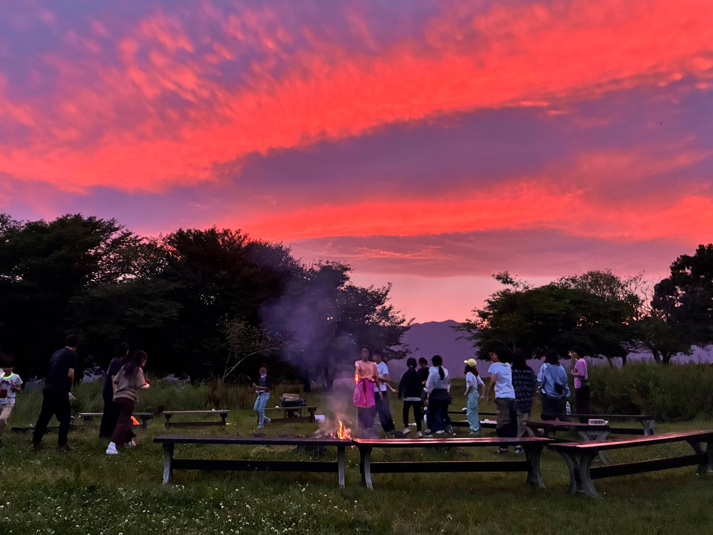 真っ赤に染まった夕焼け空の下、キャンプファイヤーを囲んで集まるaini schoolの生徒とスタッフたち