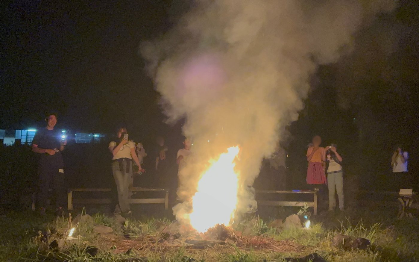 夜空の下、キャンプファイヤーに花火を入れた大きな炎と煙を囲んで見守るaini schoolの参加者たち
