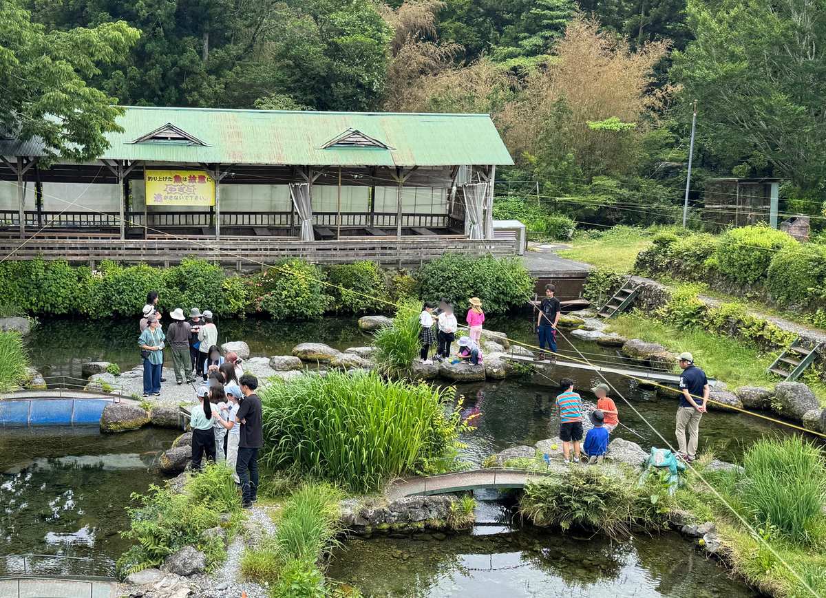 緑に囲まれた釣り堀の池の全景、生徒とスタッフたちが各所で竿を垂らす様子
