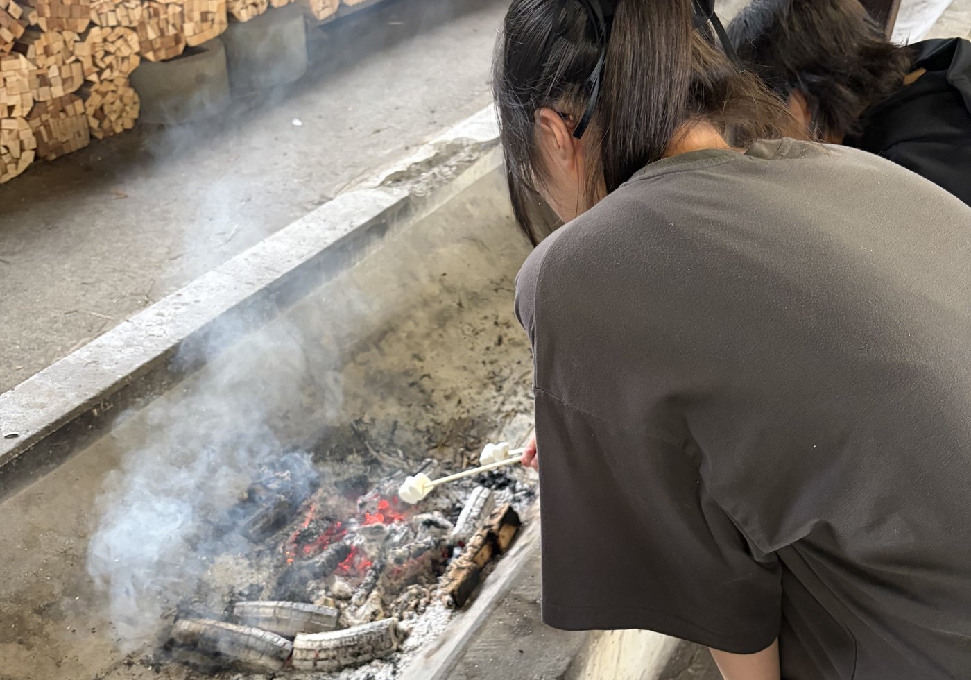 炭火の炉にマシュマロをかざして焼く生徒の後ろ姿、煙が漂う